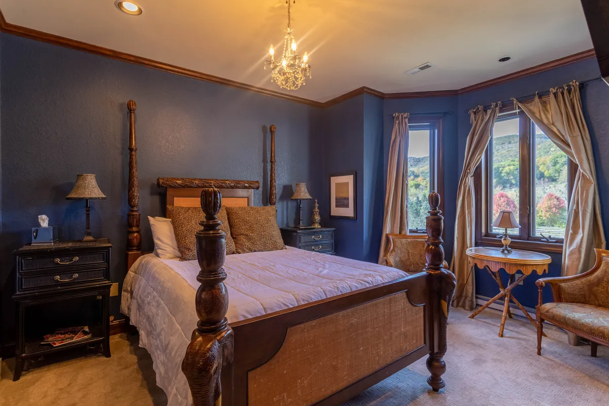 A serene bedroom with a four-poster bed, chandelier, large windows revealing beautiful outdoor scenery, a decorative armchair, and a nightstand. The setting is soothing, perfect for restful nights and quiet afternoons.
