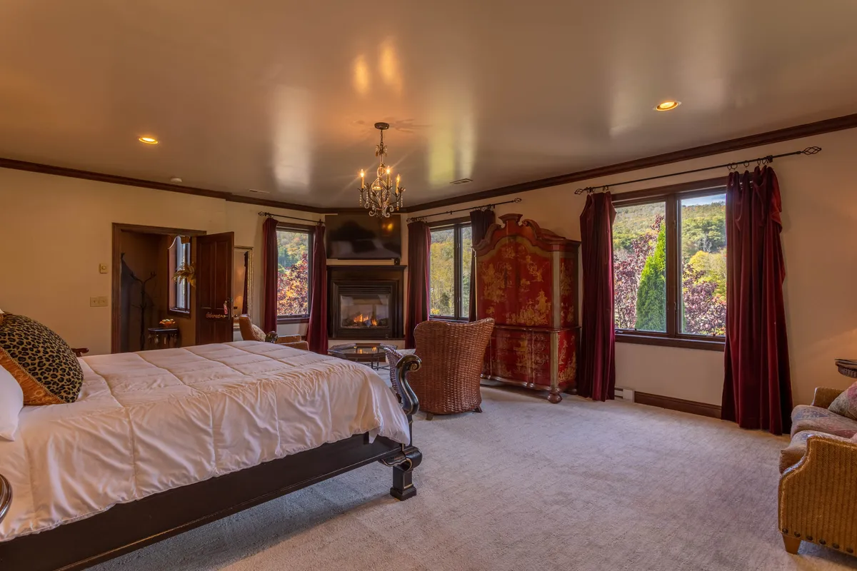 This image features a king-sized bed set in a spacious suite, complemented by a warm fireplace, large windows that offer scenic views of the surrounding landscape, an ornate wardrobe, and a comfortable seating area. The mood is tranquil and luxurious, with an emphasis on comfort and privacy.